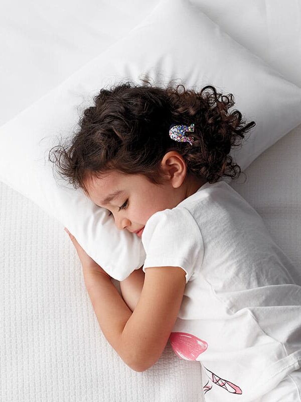 A young child with curly hair, wearing a white shirt and a hair clip, sleeps on a white pillow and bed in a peaceful posture.
