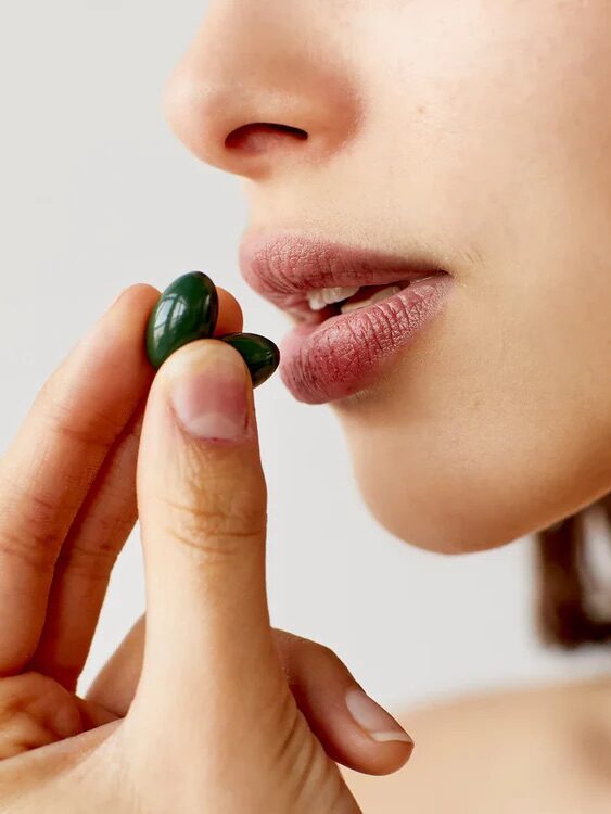 A close-up of a person's face with their mouth slightly open as they hold two green capsules near their lips with their left hand.