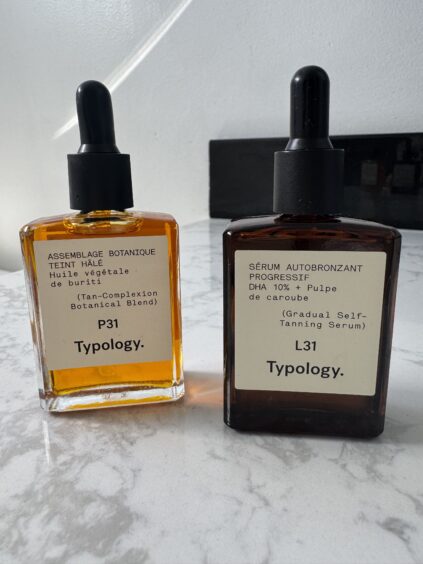 Two Typology skincare bottles on a marble surface. One bottle contains tan-complexion botanical blend oil, and the other is a gradual self-tanning serum.