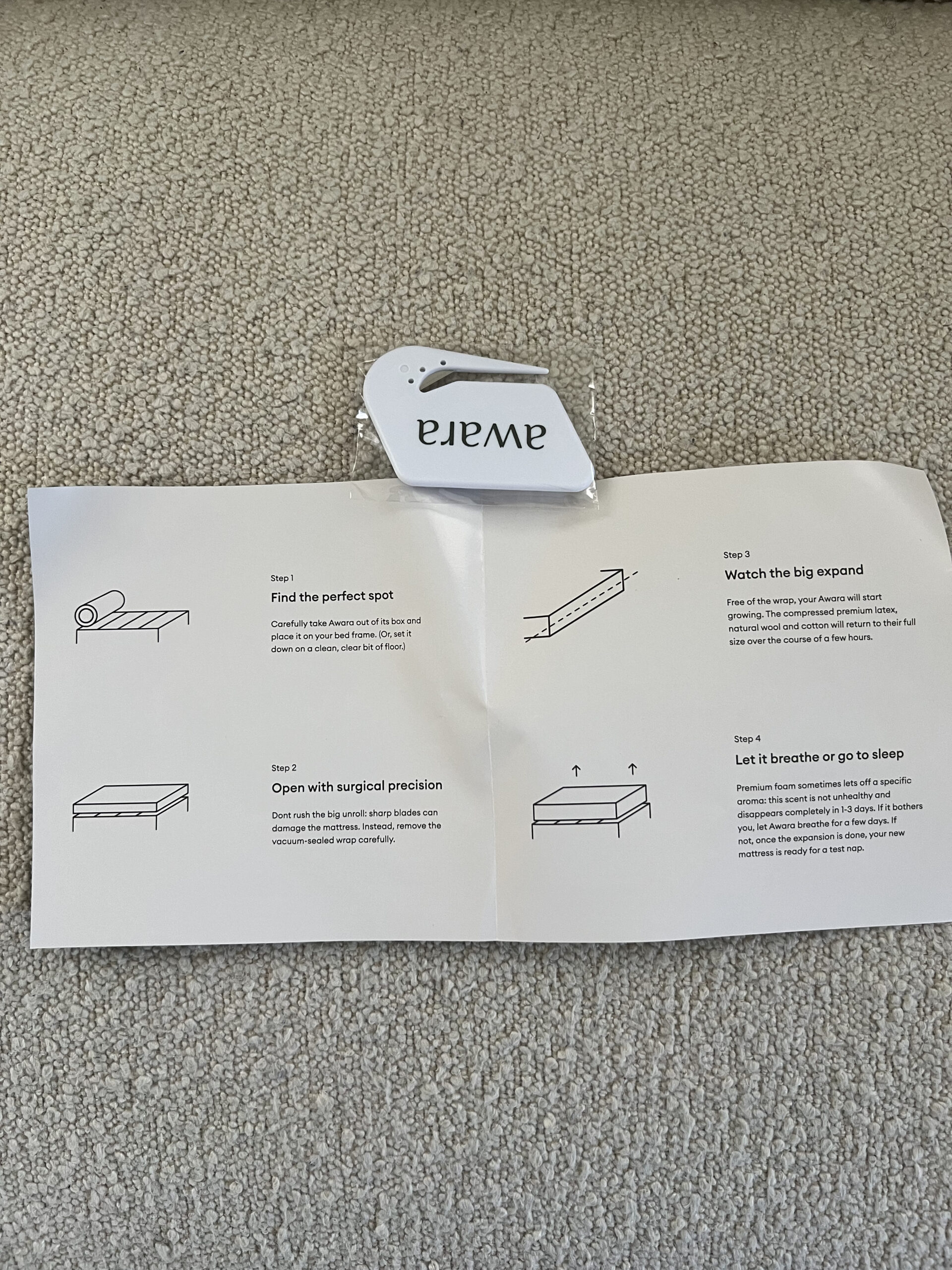 Instruction manual on a beige carpet with four steps for setting up a mattress: find the spot, watch it expand, open packaging, and let it breathe or use it. A white tag is placed at the top.