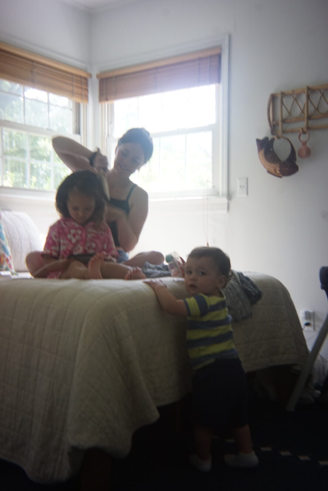 A woman is brushing the hair of a seated child on a bed, while another child stands beside the bed. The room has white walls and windows with blinds.