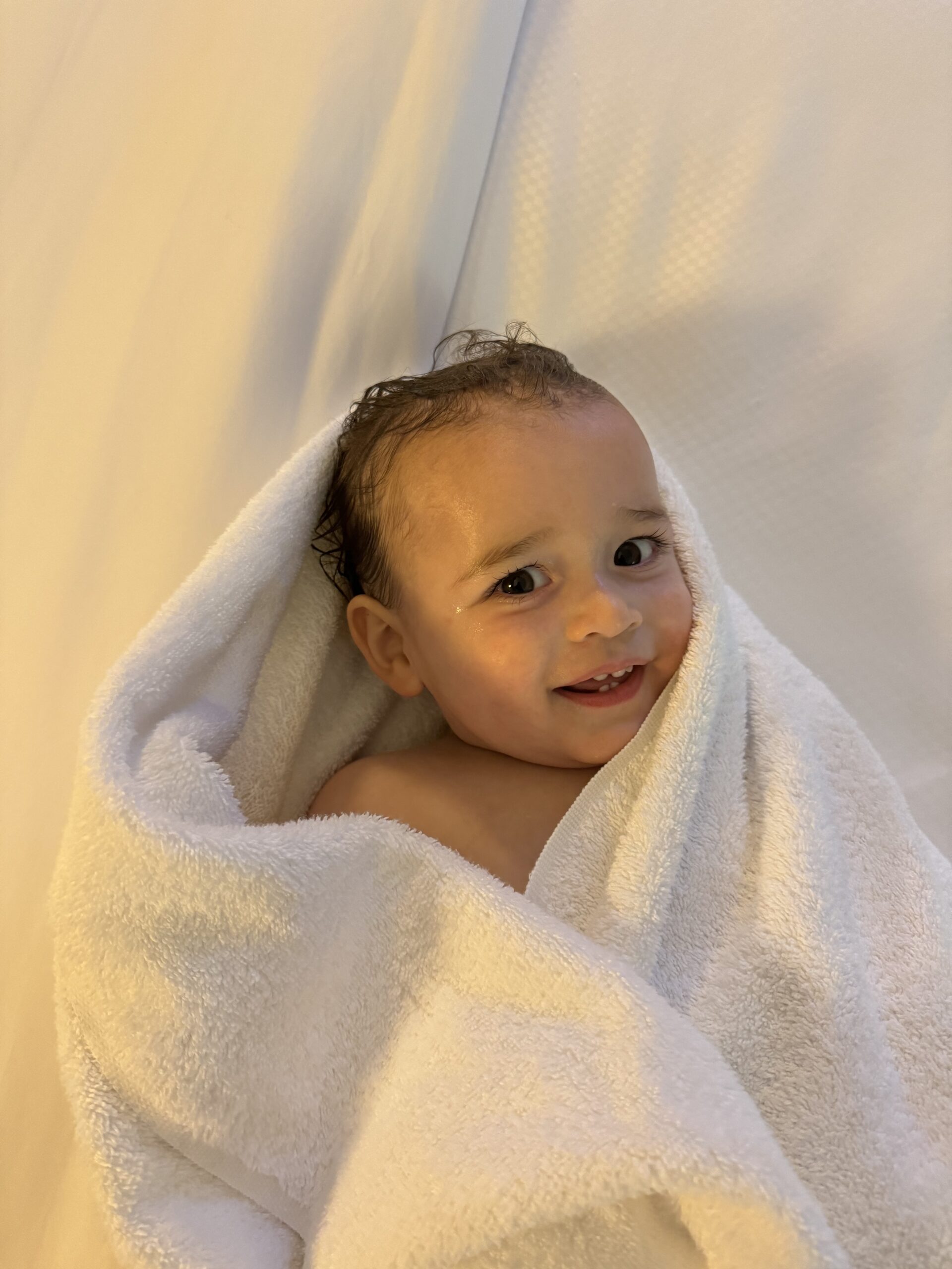 A smiling baby is wrapped in a white towel while lying on a soft surface.