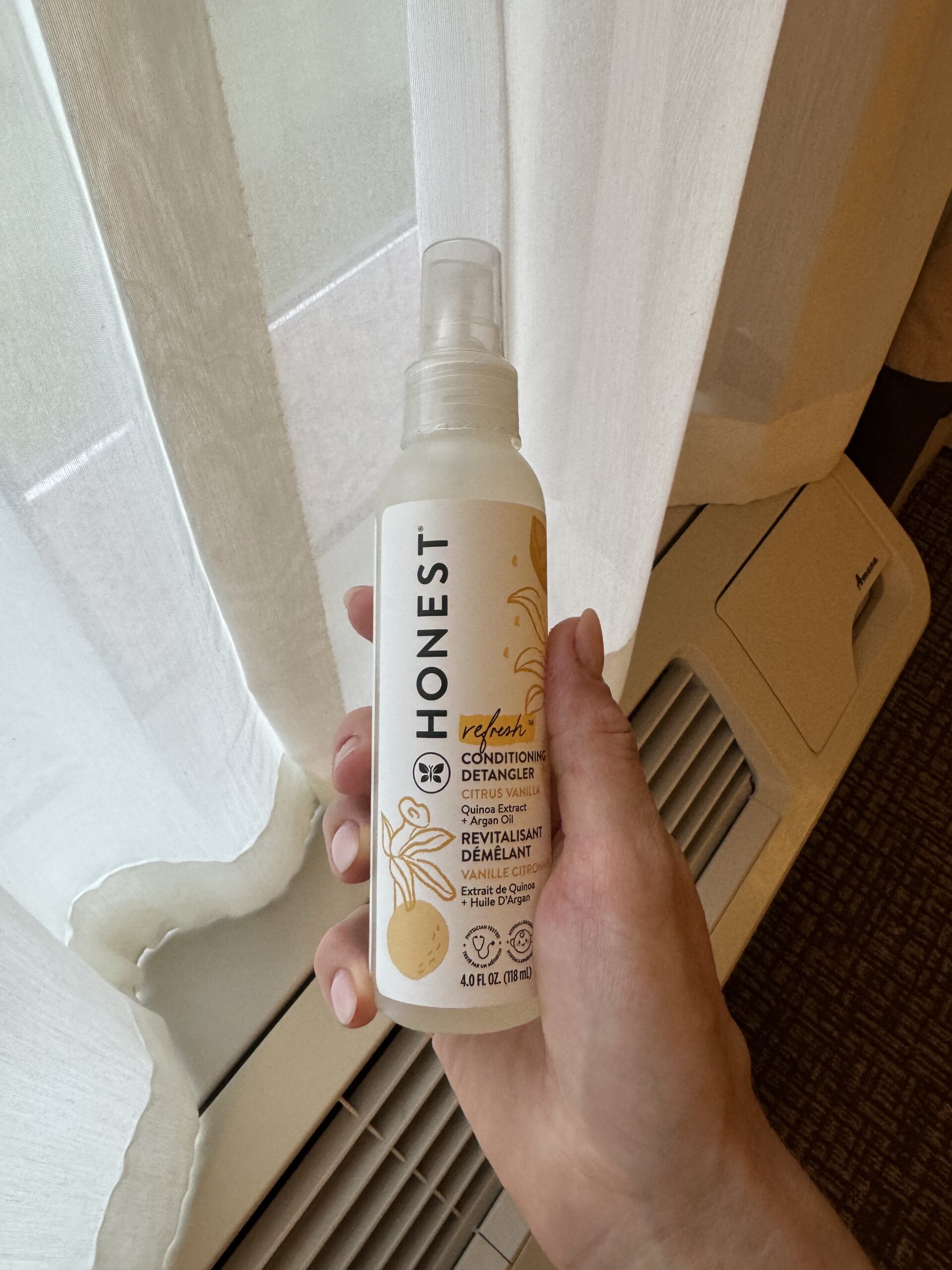 A hand holding a bottle of Honest brand conditioning detangler spray in front of a window with white curtains and an air conditioning unit below.
