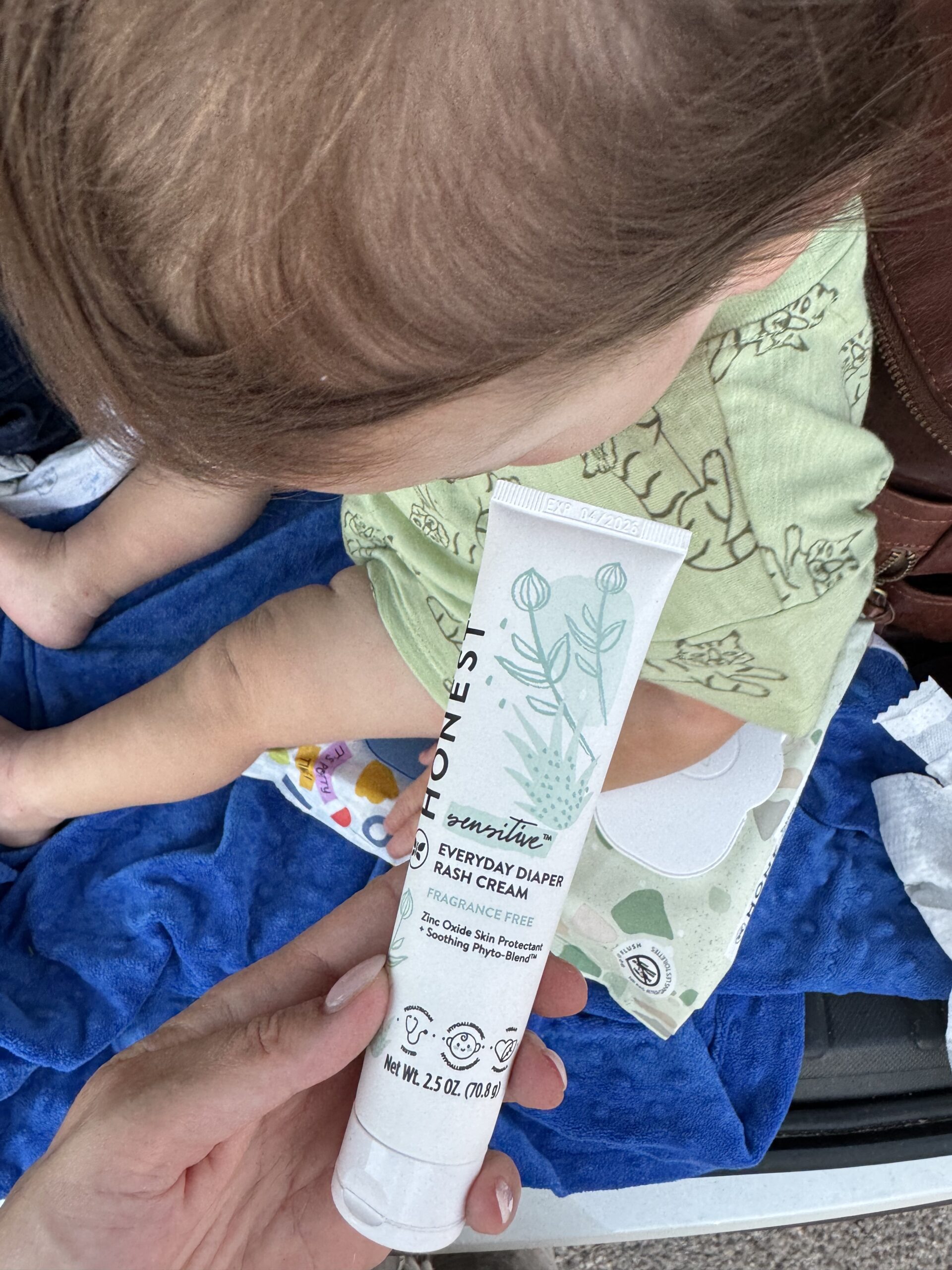 A person is holding a tube of Everyday Diaper Rash Cream next to a child wearing a green outfit, with a blue blanket and baby wipes visible in the background.