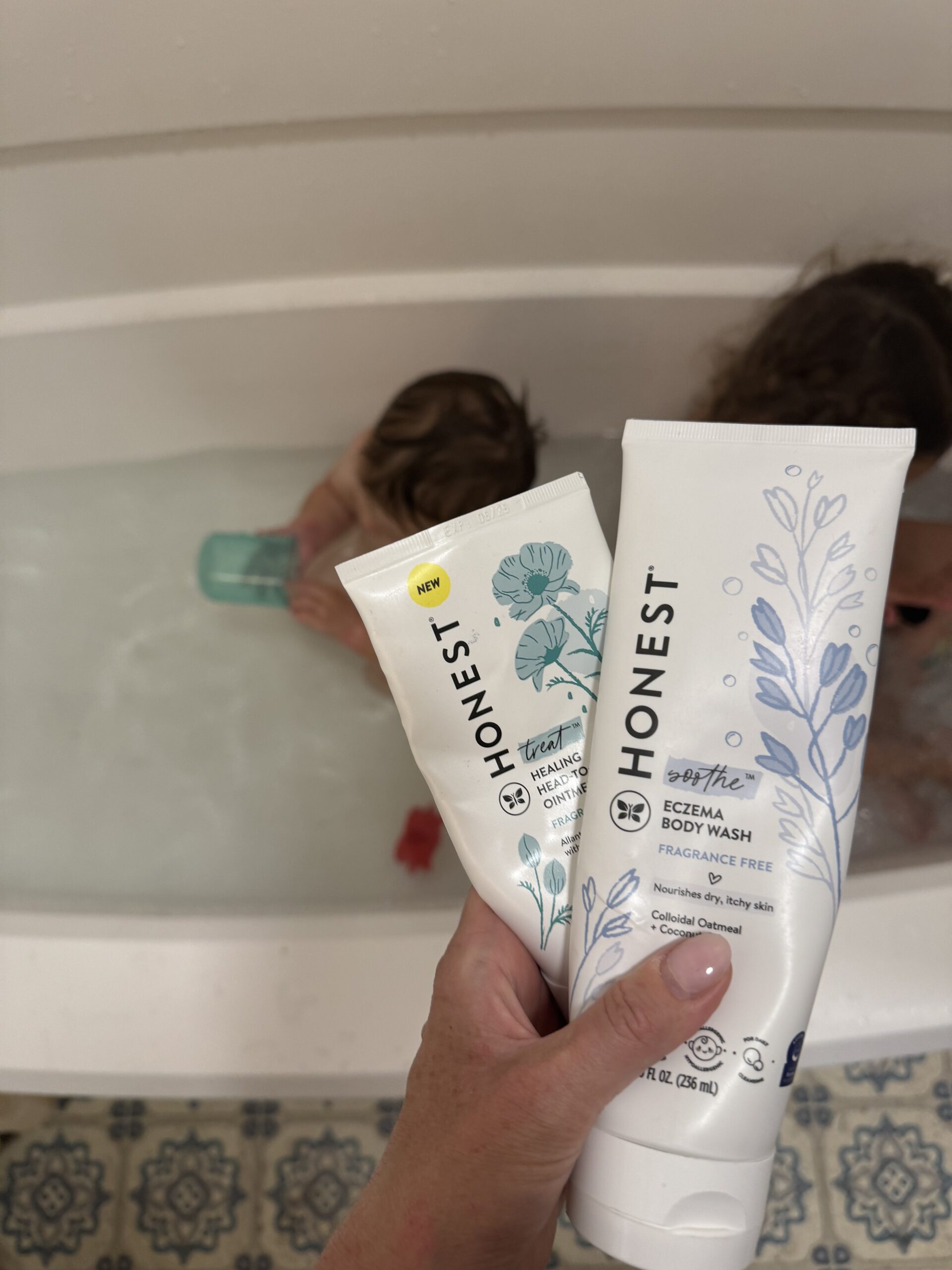 A person holds two Honest brand skincare products near a bathtub where two children are playing with bath toys.