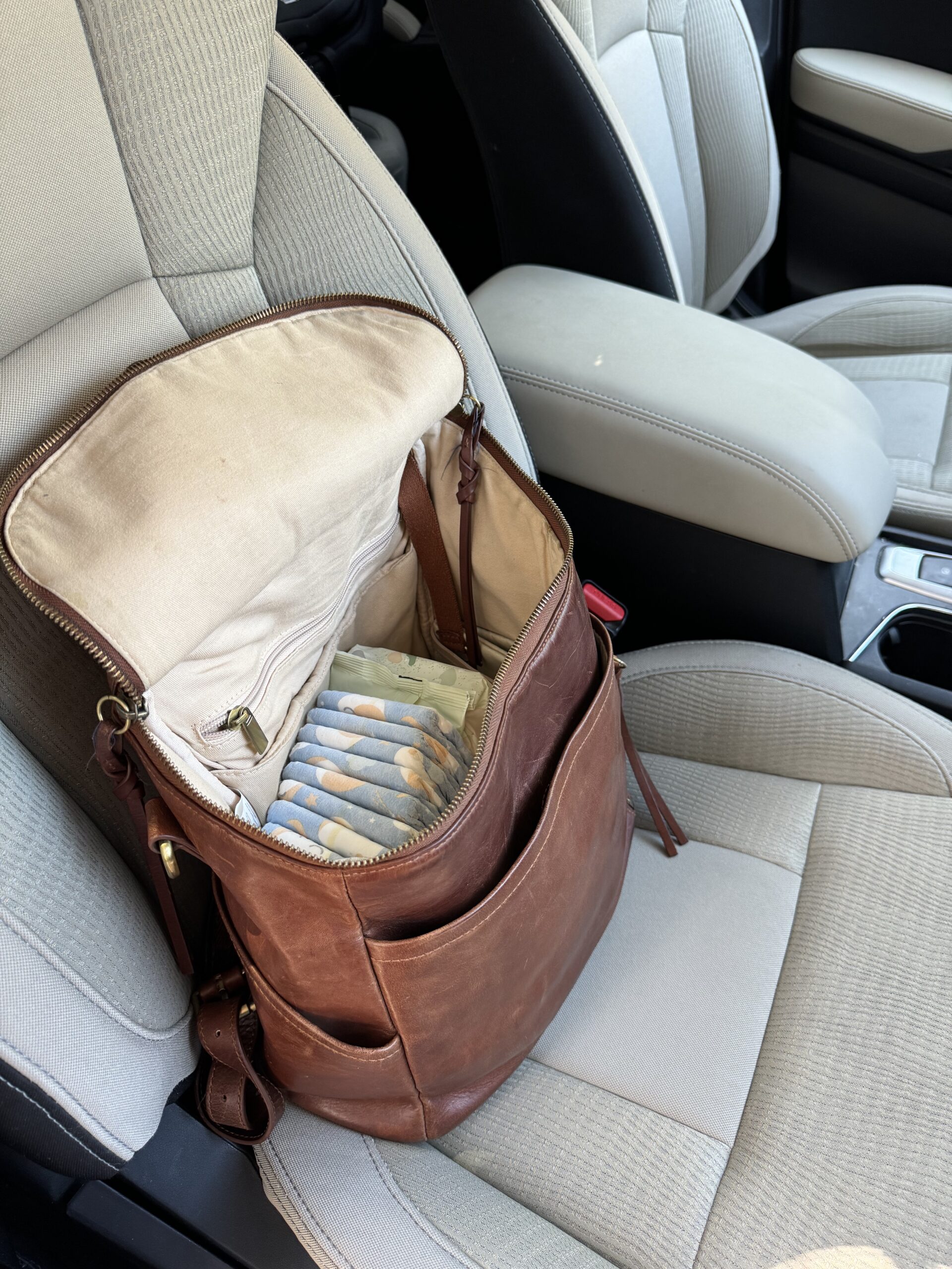 An open brown leather backpack with diapers inside is placed on the passenger seat of a car.