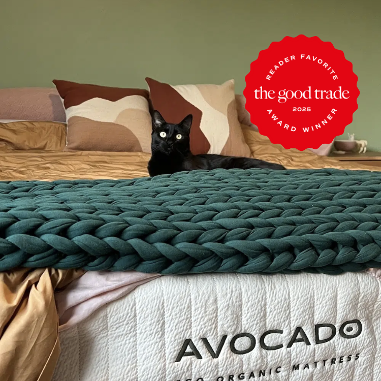 A black cat sits on a bed with a chunky green blanket. A red award badge for "The Good Trade Reader Favorite 2025" is displayed on the right. The mattress label reads "Avocado.