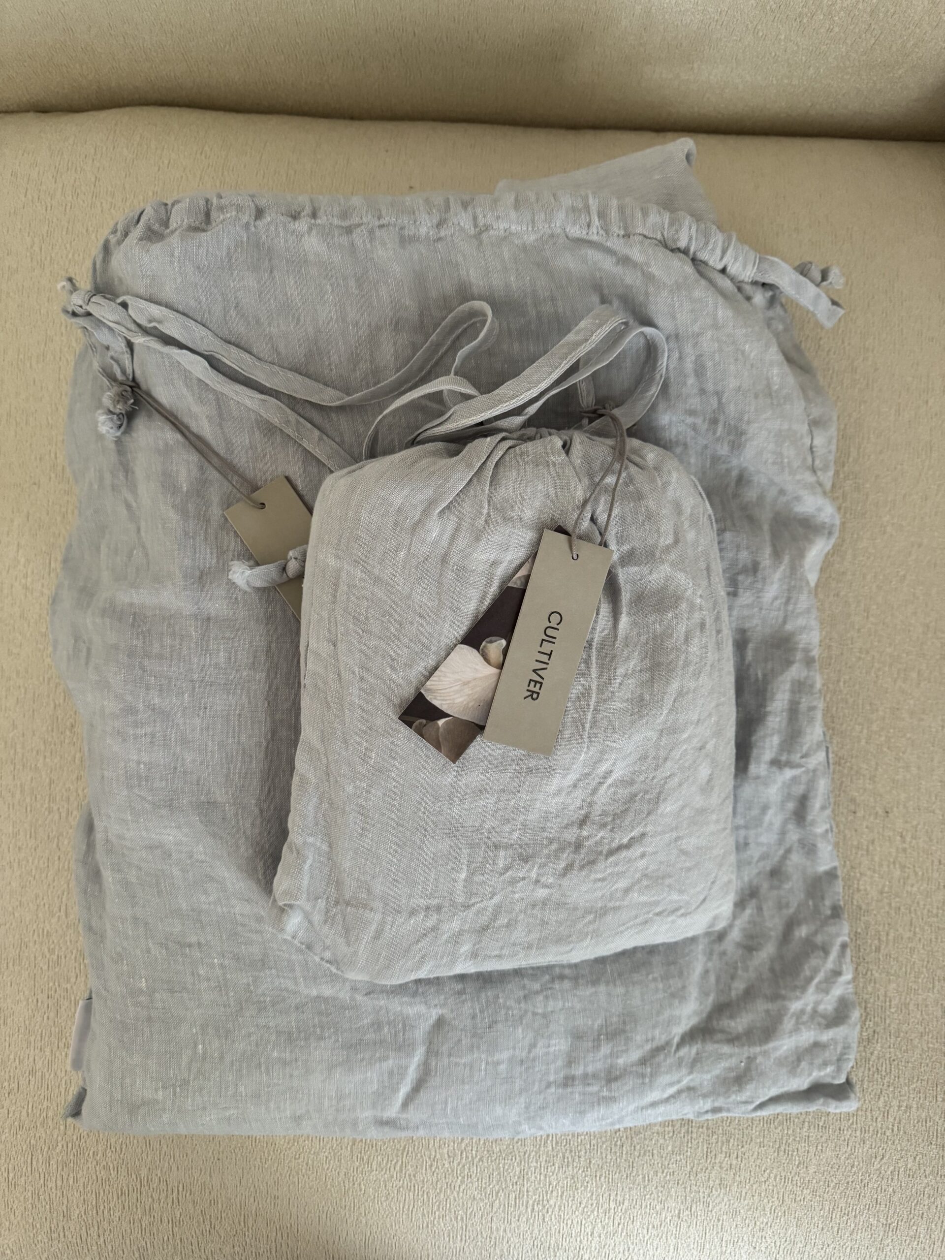 Two gray, drawstring linen bags with tags attached, placed on a light-colored surface.