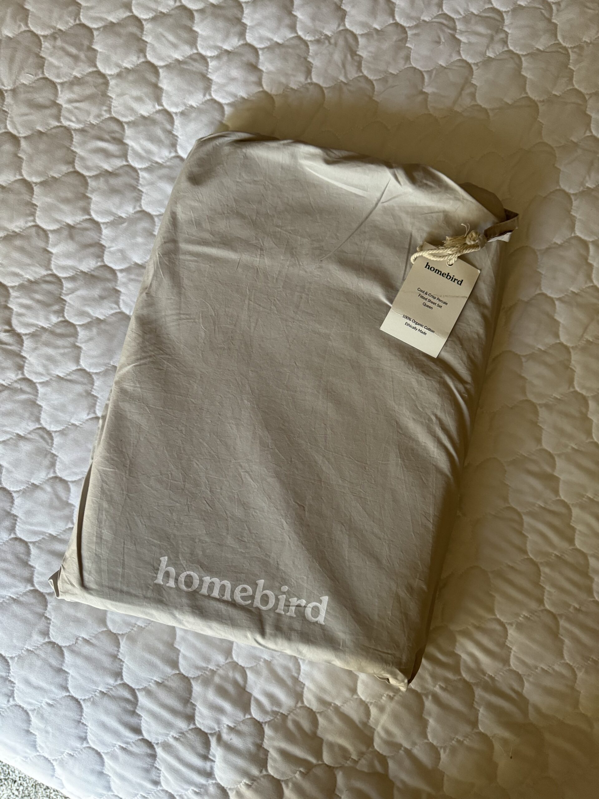 A packaged set of Homebird bed sheets with a label attached, placed on a white quilted surface.