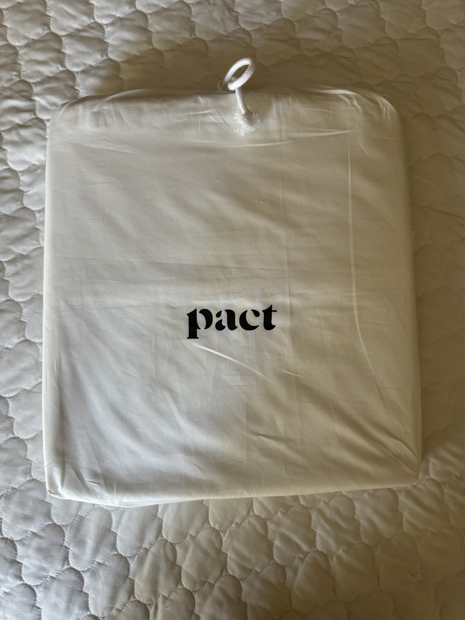 A sealed white package labeled "pact" rests on a quilted surface, with a small loop at the top.