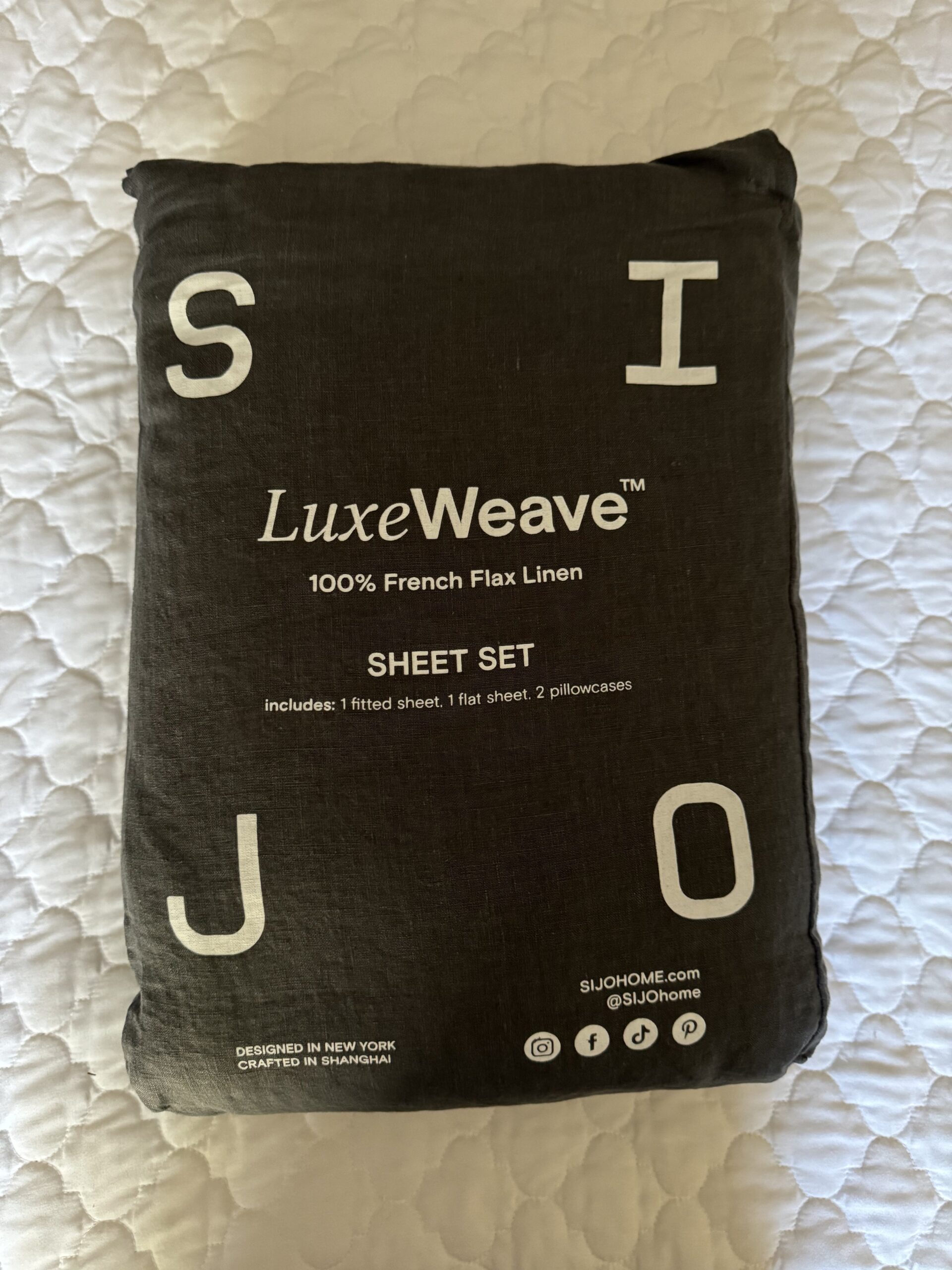 A packaged LuxeWeave sheet set made of 100% French Flax Linen sits on a quilted surface. The package includes one fitted sheet, one flat sheet, and two pillowcases.