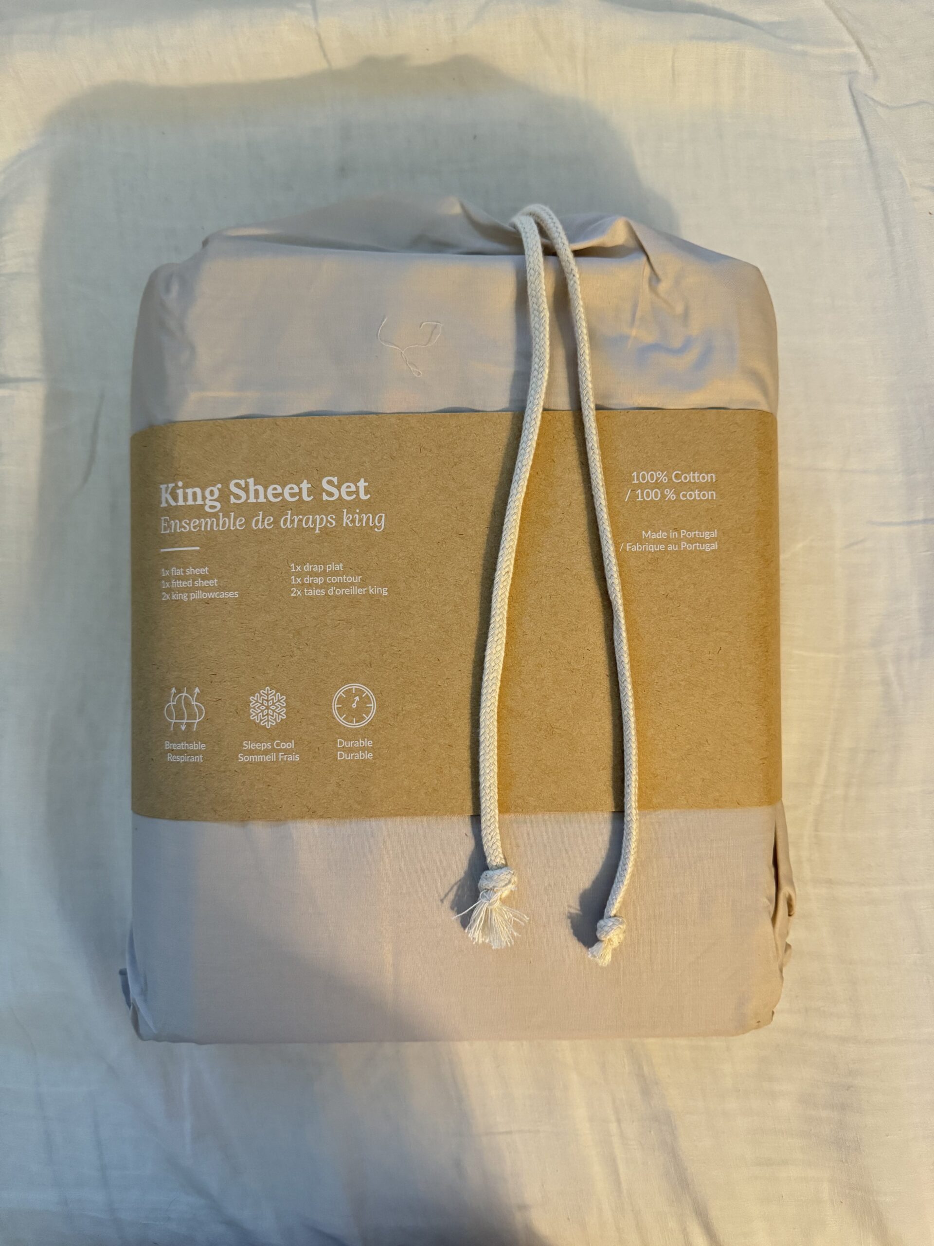 Packaged king sheet set with a cardboard label detailing product features and material, tied with white drawstrings. The sheet set is described as 100% cotton and made in Portugal.