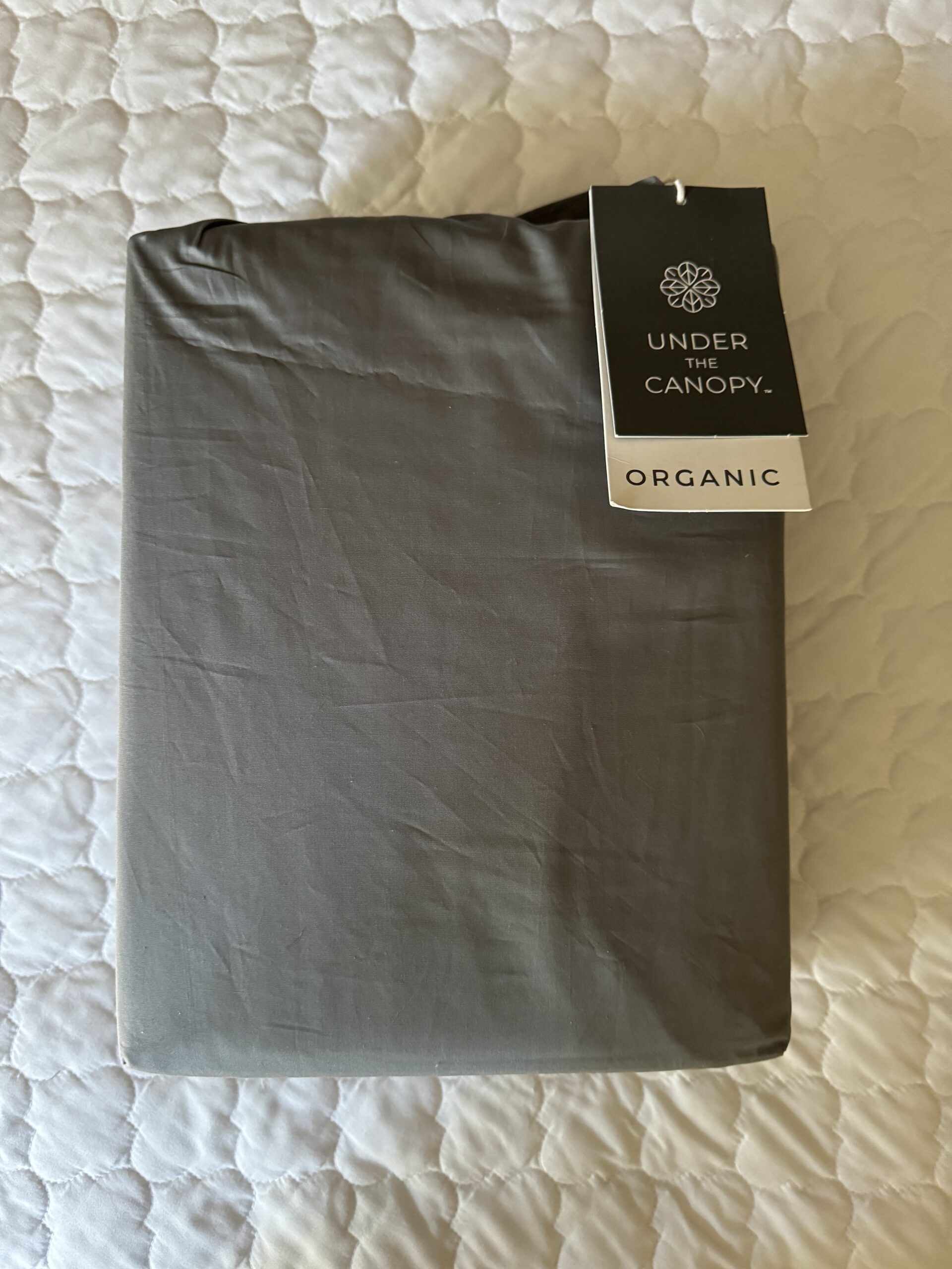 A neatly folded gray sheet with an attached tag labeled "Under the Canopy" and "Organic," resting on a quilted white surface.