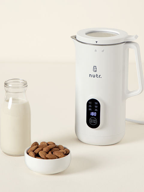 A white electric nut milk maker with digital display is placed beside a small bottle filled with milk, and a bowl of almonds.