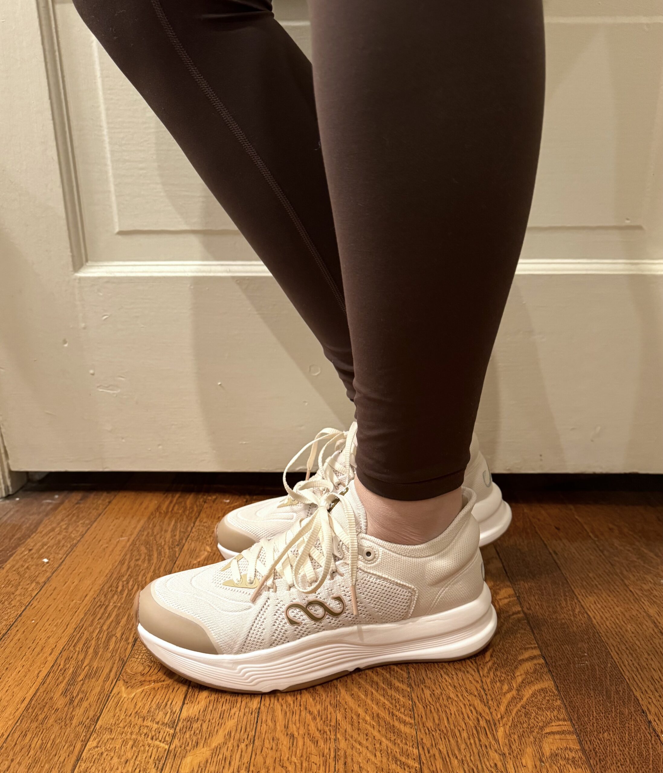 Person wearing white sneakers and dark leggings stands on a wooden floor near a door.