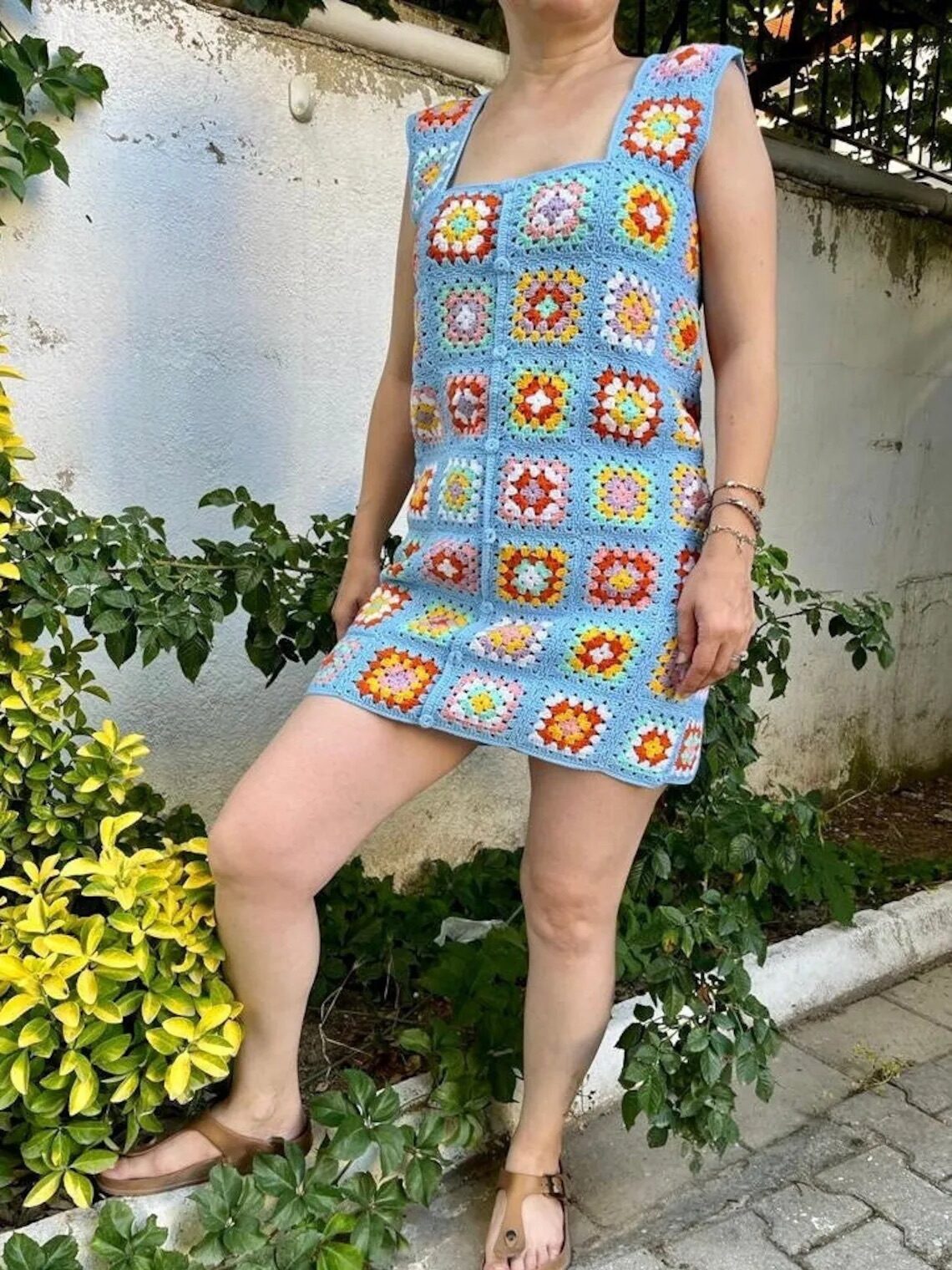A person wearing a sleeveless dress made of colorful granny squares stands outside near green and yellow foliage, with one leg bent slightly forward.
