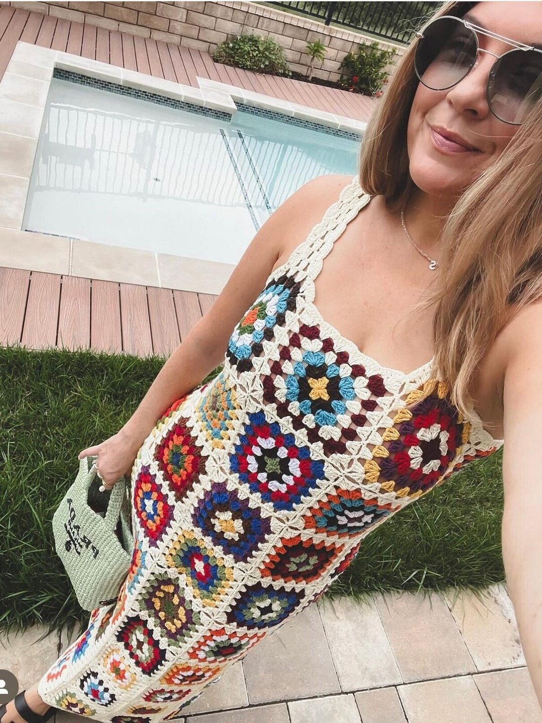 A woman wearing sunglasses and a colorful crocheted dress stands near a pool, holding a green bag.