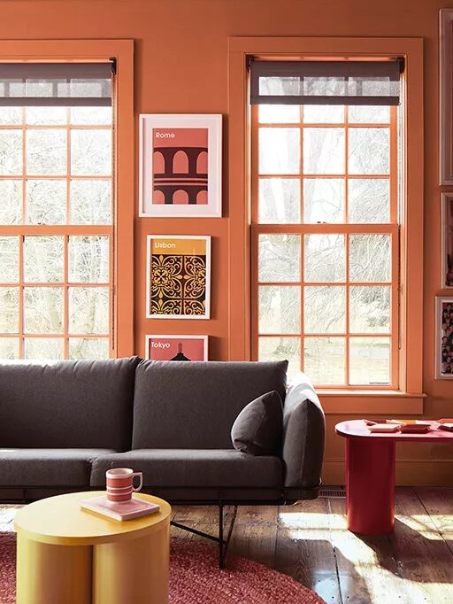A room with orange walls, two large windows, a gray couch, and three framed pictures. There is a yellow circular table and a red side table.