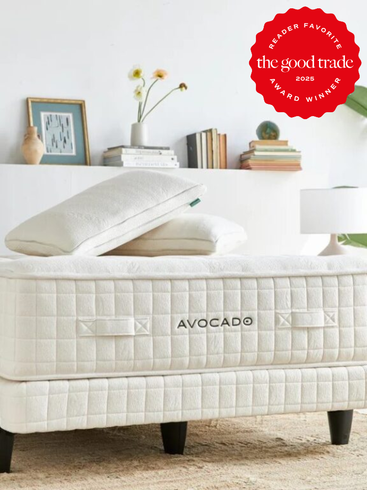 Avocado mattress and pillows on a bed frame, with a "Reader Favorite" award badge from The Good Trade. Room has minimal decor with plants and books.