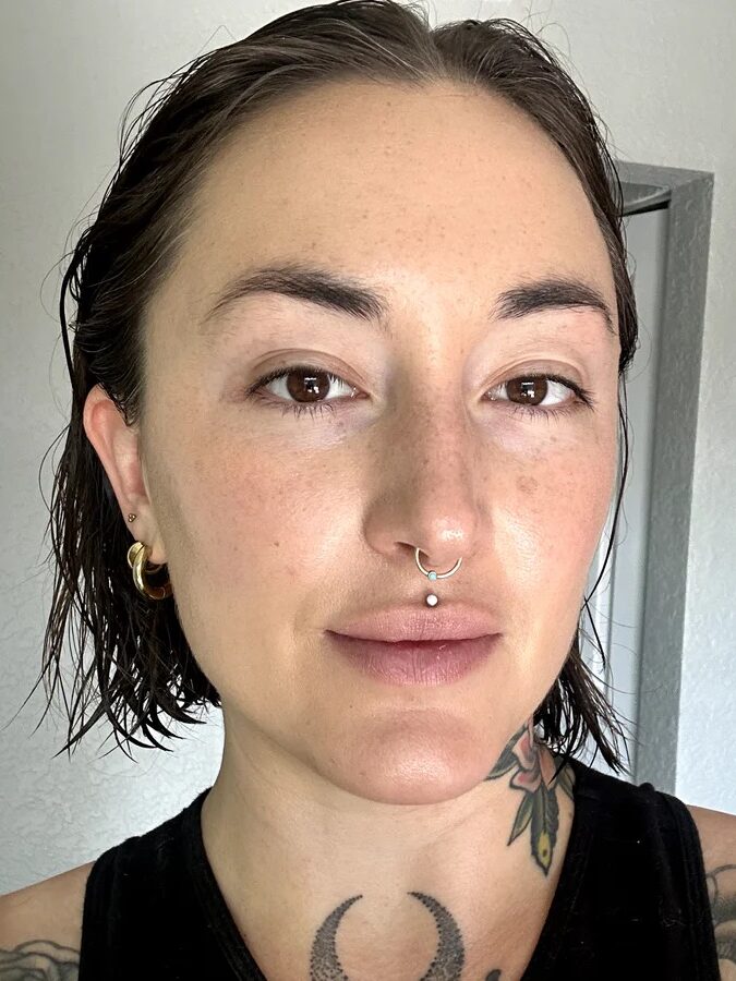 A person with short wet hair, a septum piercing, and visible tattoos stands indoors, looking directly at the camera. They are wearing a black top and gold hoop earrings.