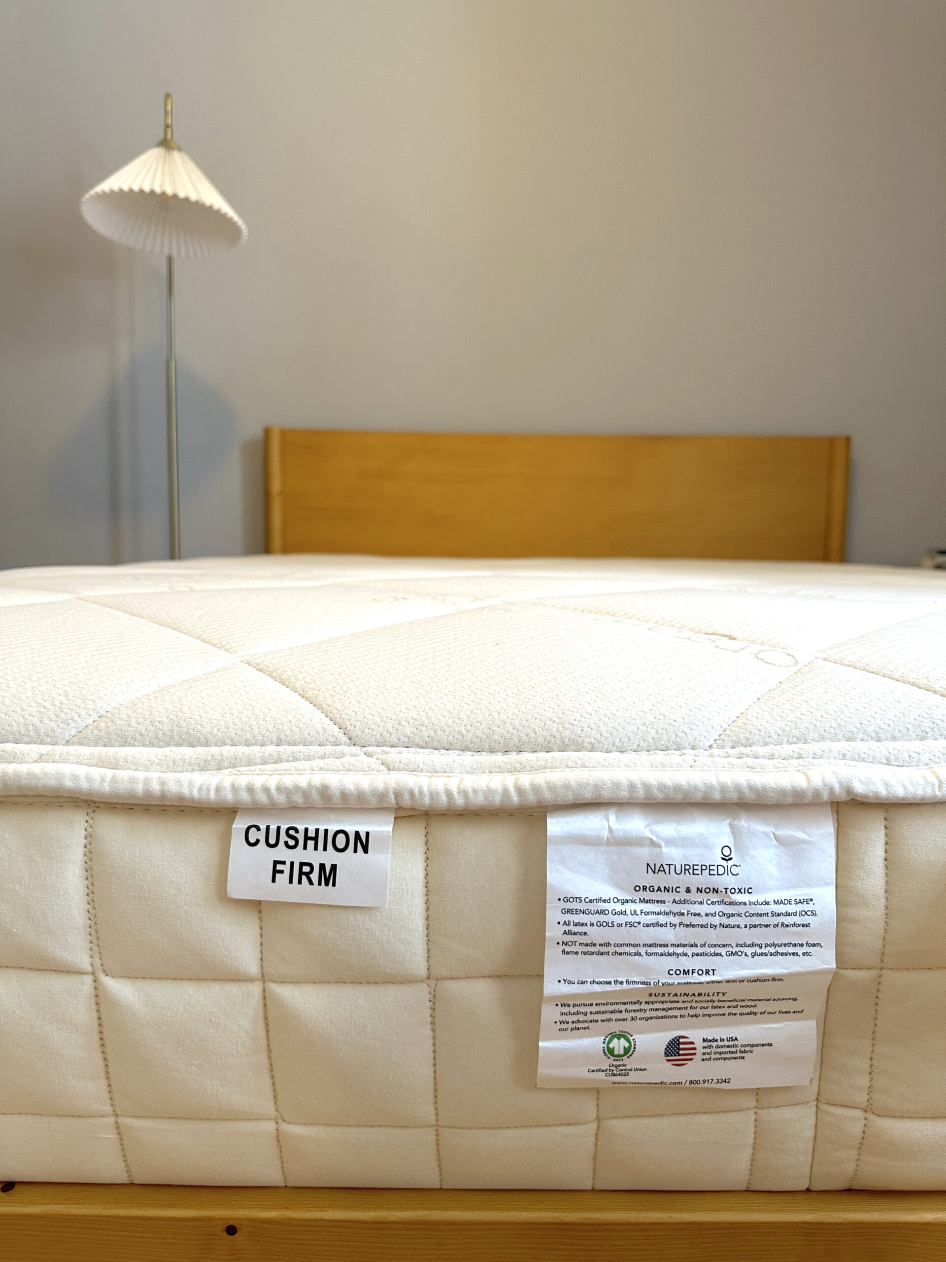 A close-up of a white mattress with "CUSHION FIRM" and product information tags, on a wooden bed frame in a room with a lamp and light gray wall.