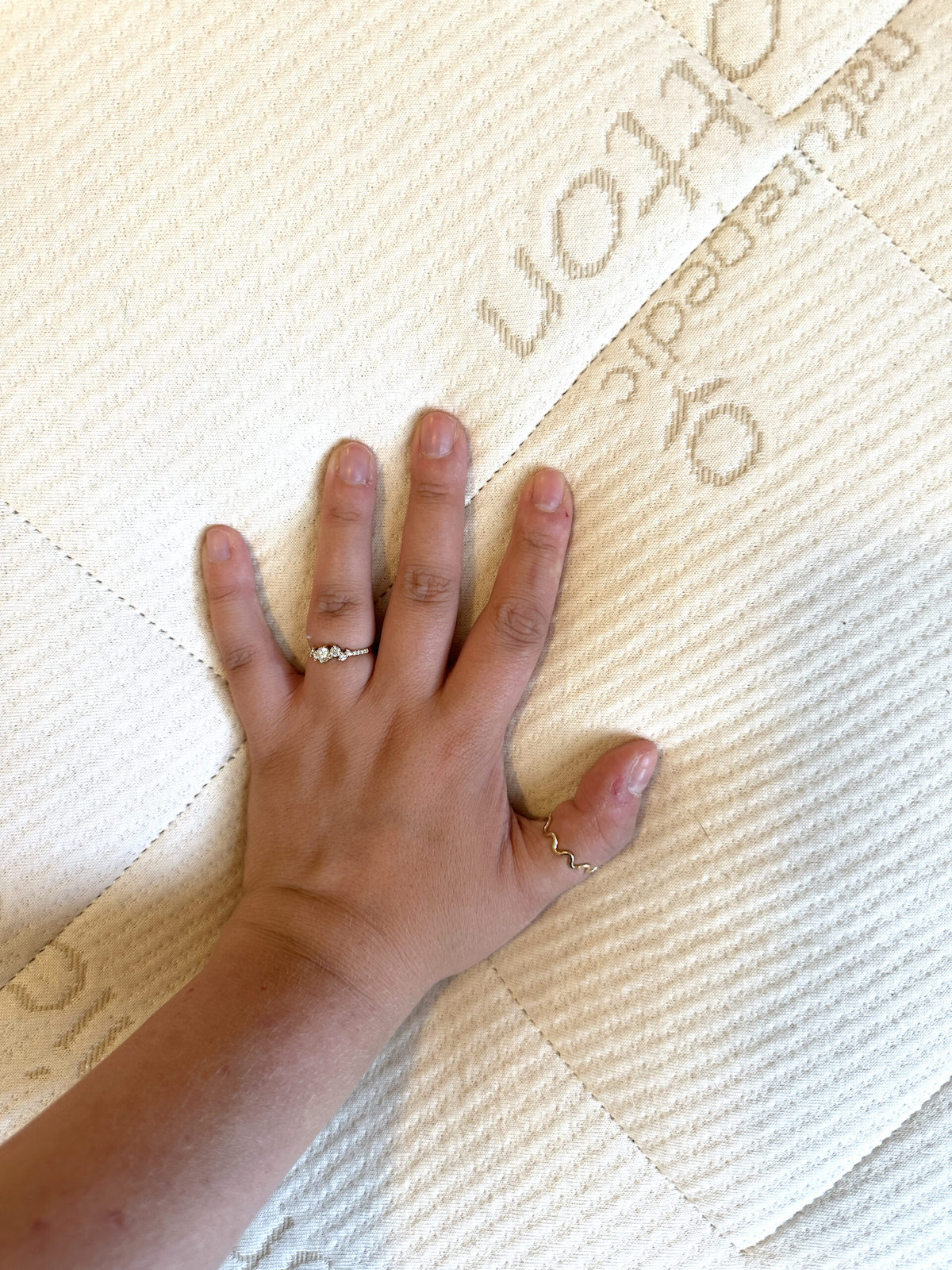 Reviewer Brianna Schubert's hand with two rings rests on a textured fabric topper of a Naturepedic organic cotton mattress.