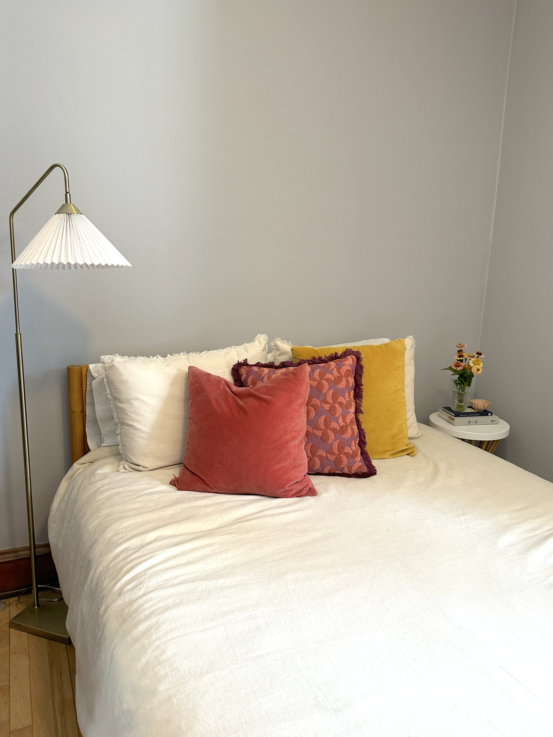 A neatly made bed with white bedding, colorful accent pillows, a floor lamp, and a small round side table with a vase of flowers.
