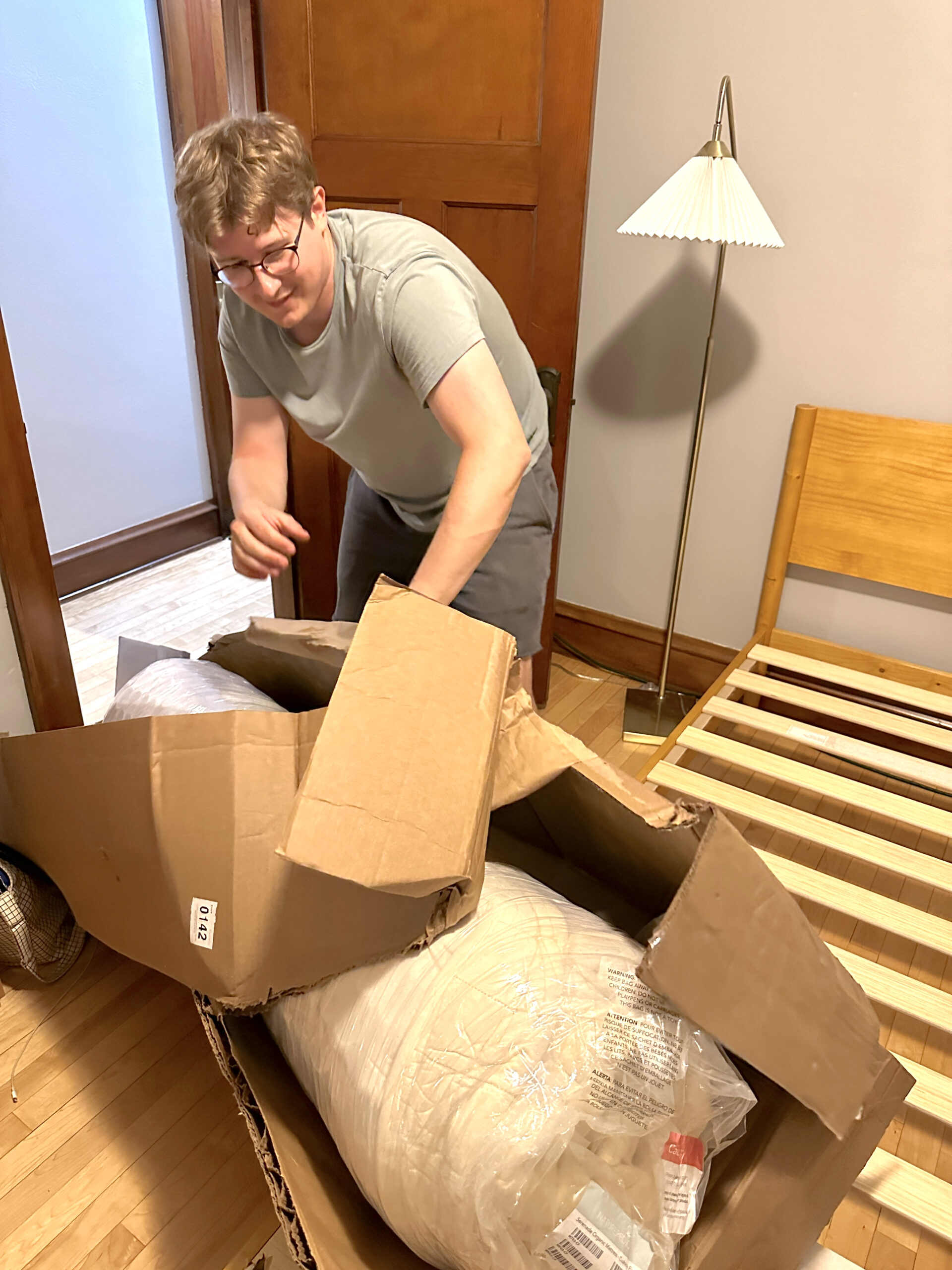 A person opens a large, partially damaged cardboard box containing a rolled-up mattress next to a wooden bed frame in a bedroom.