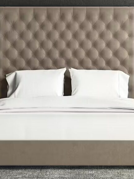 A bedroom with a large tufted headboard, a neatly made bed with white linens, two bedside tables with books, and modern lamps. Dark curtains and a city view can be seen in the background.