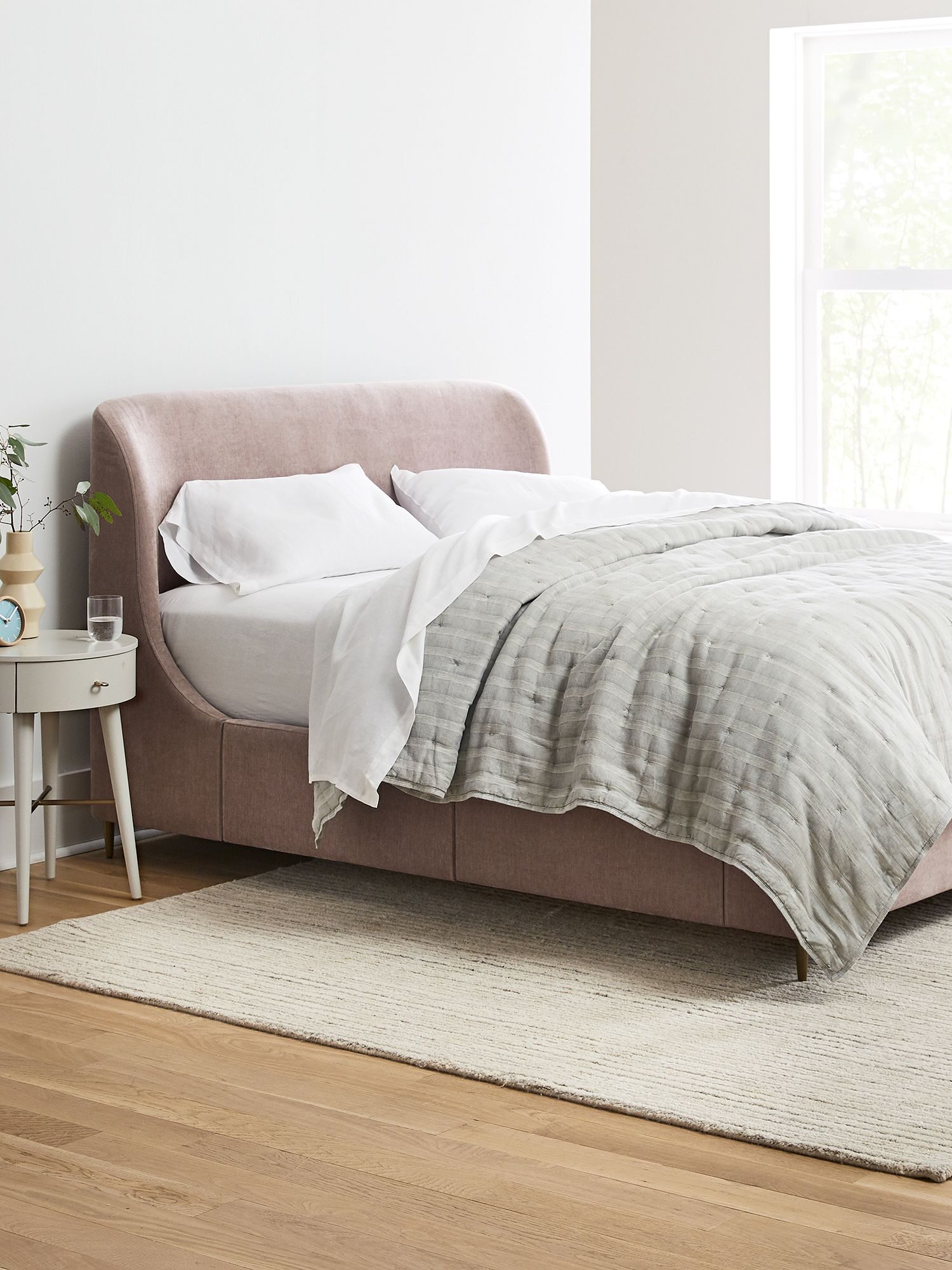 A neatly made bed with light-colored bedding, positioned on a light-colored rug in a minimalist bedroom with a window, nightstand, and dresser.