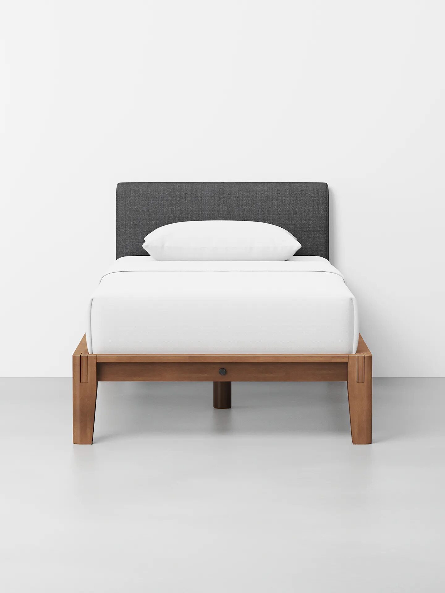 A minimalist single bed with a dark gray upholstered headboard, white bedding, and a light wooden frame against a plain white wall.