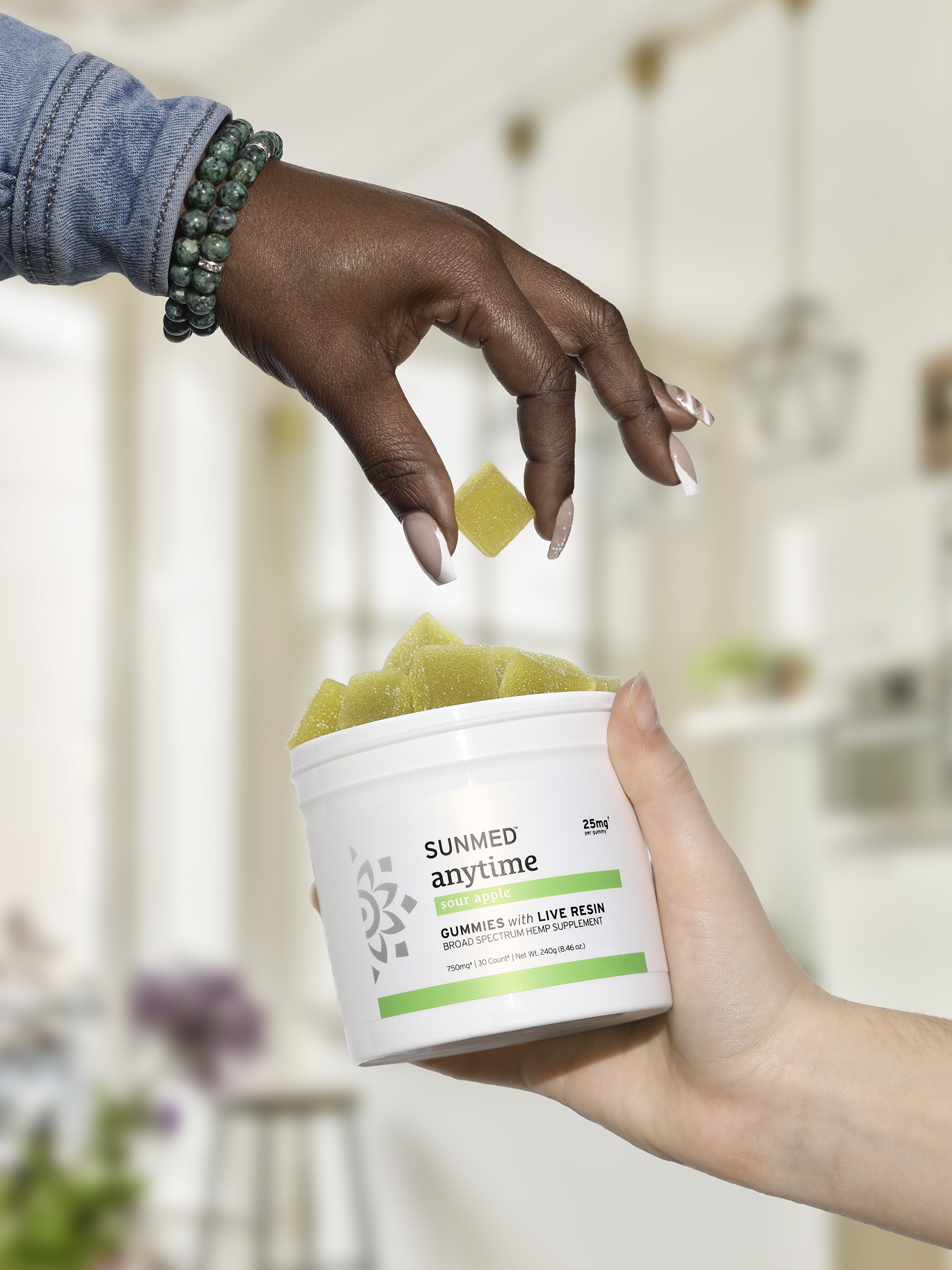 Two hands holding and reaching for a gummy from a container labeled "Sunmed Anytime Gummies with Live Resin." One hand wears a beaded bracelet. The background appears to be a bright, modern kitchen.