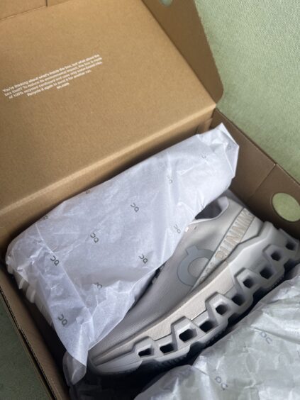A single light gray athletic shoe partially wrapped in branded tissue paper inside an open cardboard shoe box.