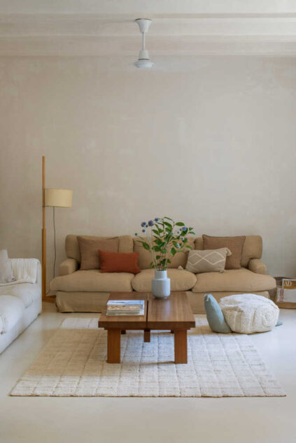 A neutral-toned living room with a beige sofa, wooden coffee table, pouf, floor lamp, and a plant in a vase on the table, set on a light rug.