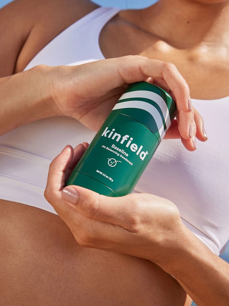 Person in white clothing holding a green "Kinfield" branded spray bottle against the background of their torso.