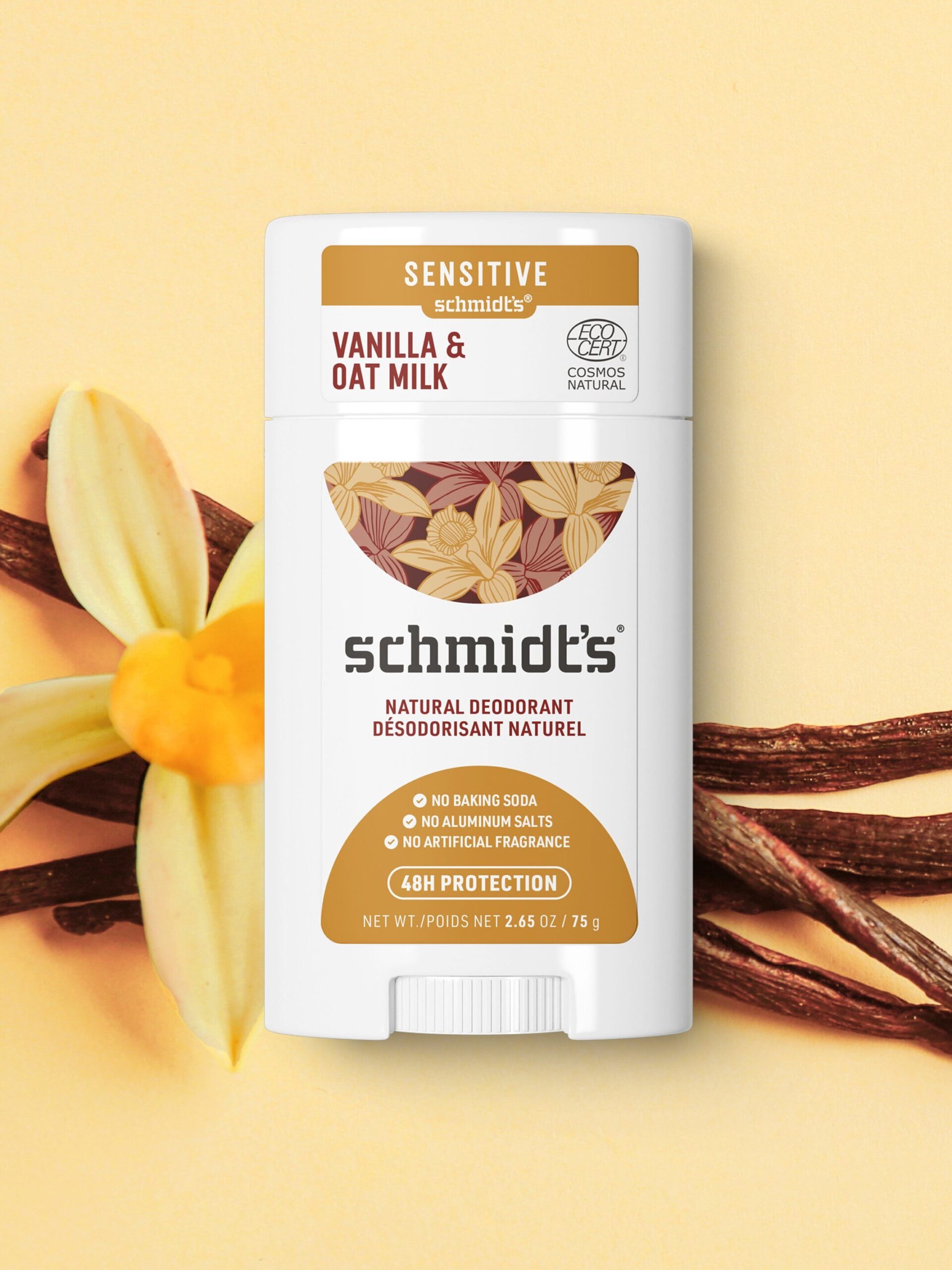 A stick of Schmidt's Vanilla and Oat Milk natural deodorant is placed against a light background with vanilla flowers and beans. The label highlights its sensitive formula and natural ingredients.