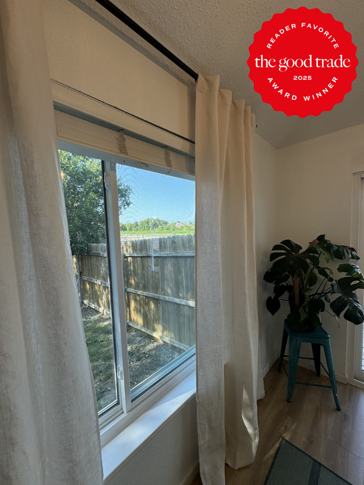 Room with a window covered by white curtains, a wooden floor, and a plant on a green stool. There's an award badge for "The Good Trade 2025" on the top right corner.