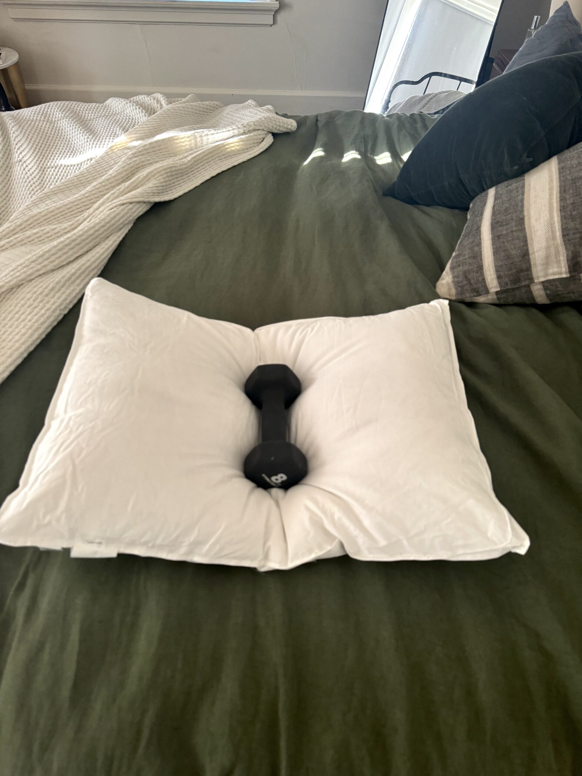 A white pillow with an indentation in the center is placed on a green bedspread. A black dumbbell rests in the middle of the pillow.