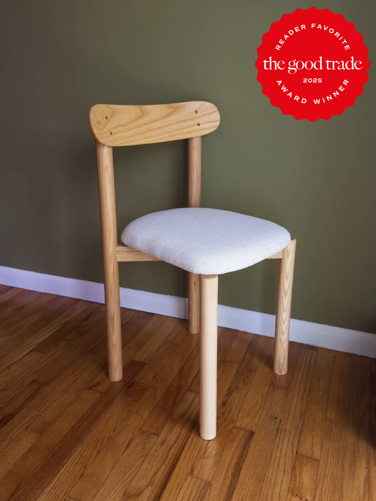 Wooden chair with a light fabric seat against a green wall on a wooden floor. A red "The Good Trade 2025 Award Winner" badge is displayed in the top right corner.
