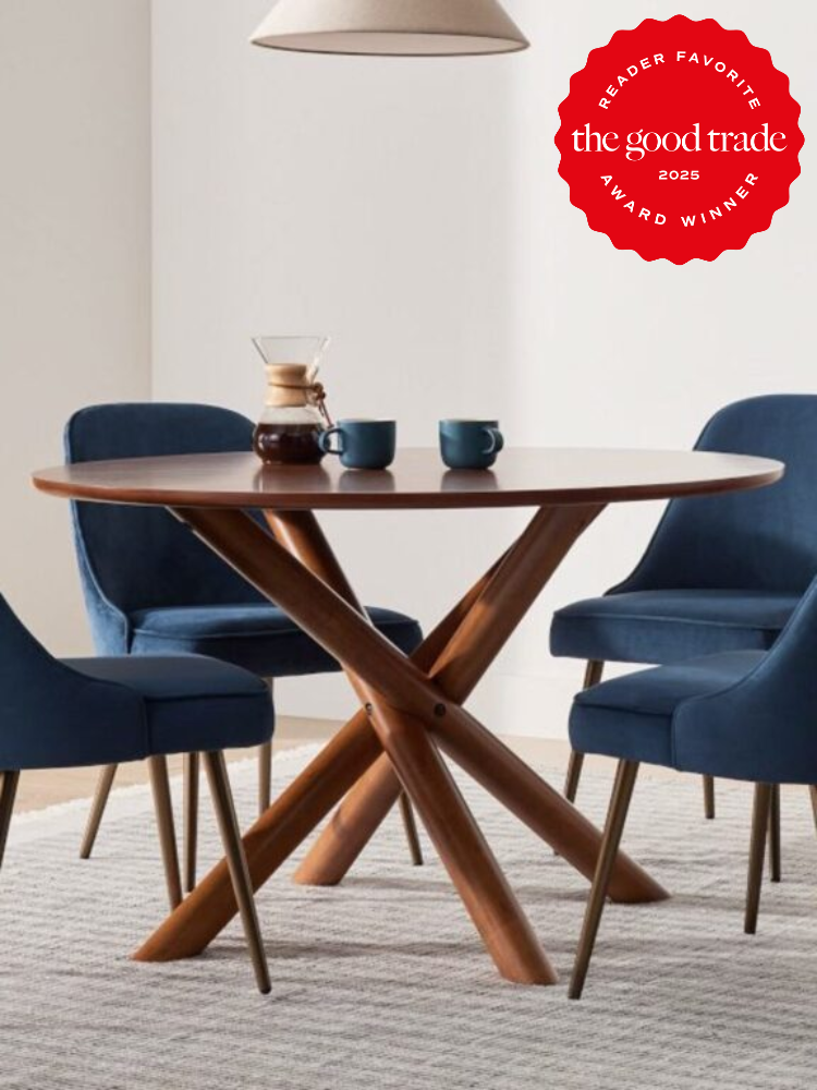 Round wooden table with crossed legs, set with a carafe and two blue mugs, surrounded by four blue upholstered chairs on a gray rug. Red award badge in the top corner reads "The Good Trade 2025.