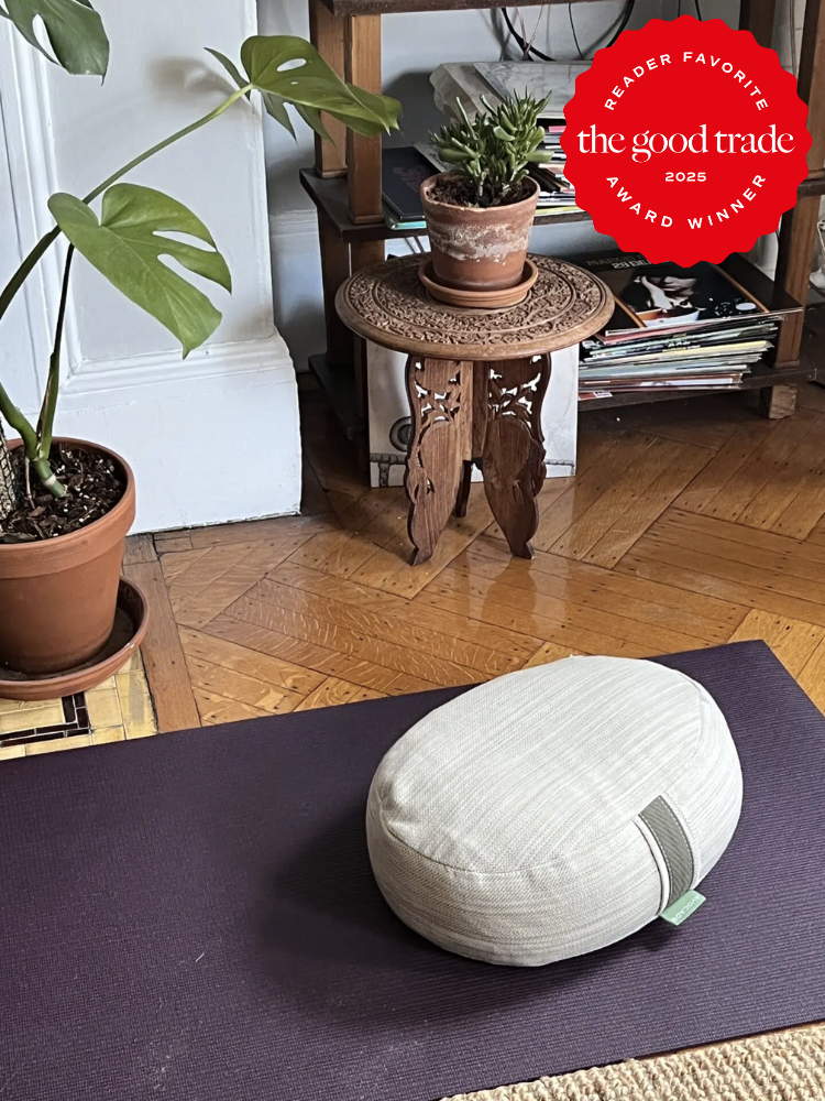 A meditation cushion on a yoga mat, next to potted plants and a small wooden table with magazines. Red sticker indicating "Reader Favorite, the good trade, award winner 2025.