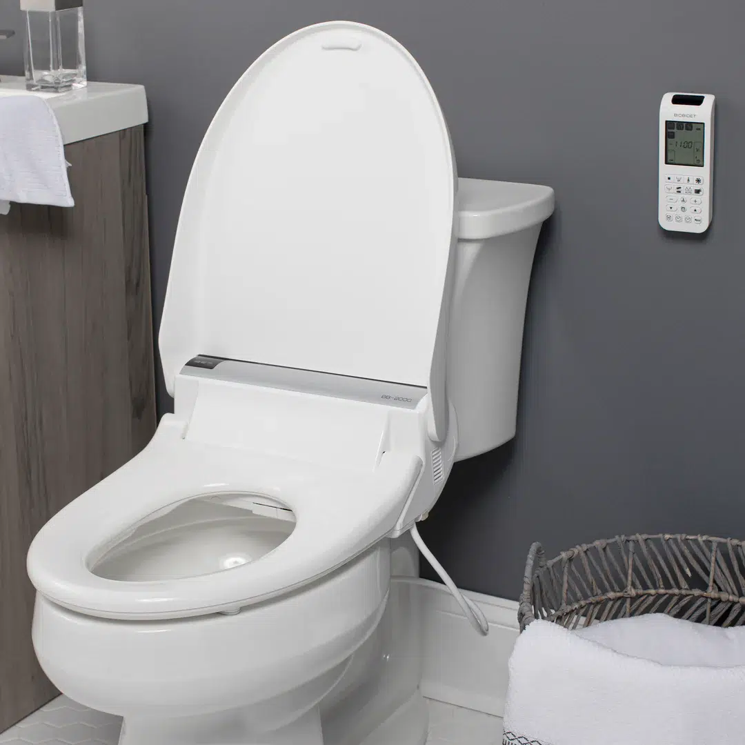 A white smart toilet with an open lid, electronic controls on the side, and a wall-mounted remote control next to a laundry basket in a modern bathroom.