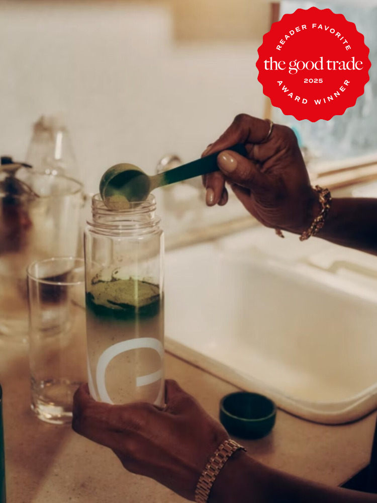 Person adding green powder to clear water bottle on kitchen counter; "the good trade 2025 award winner" badge in the top right corner.