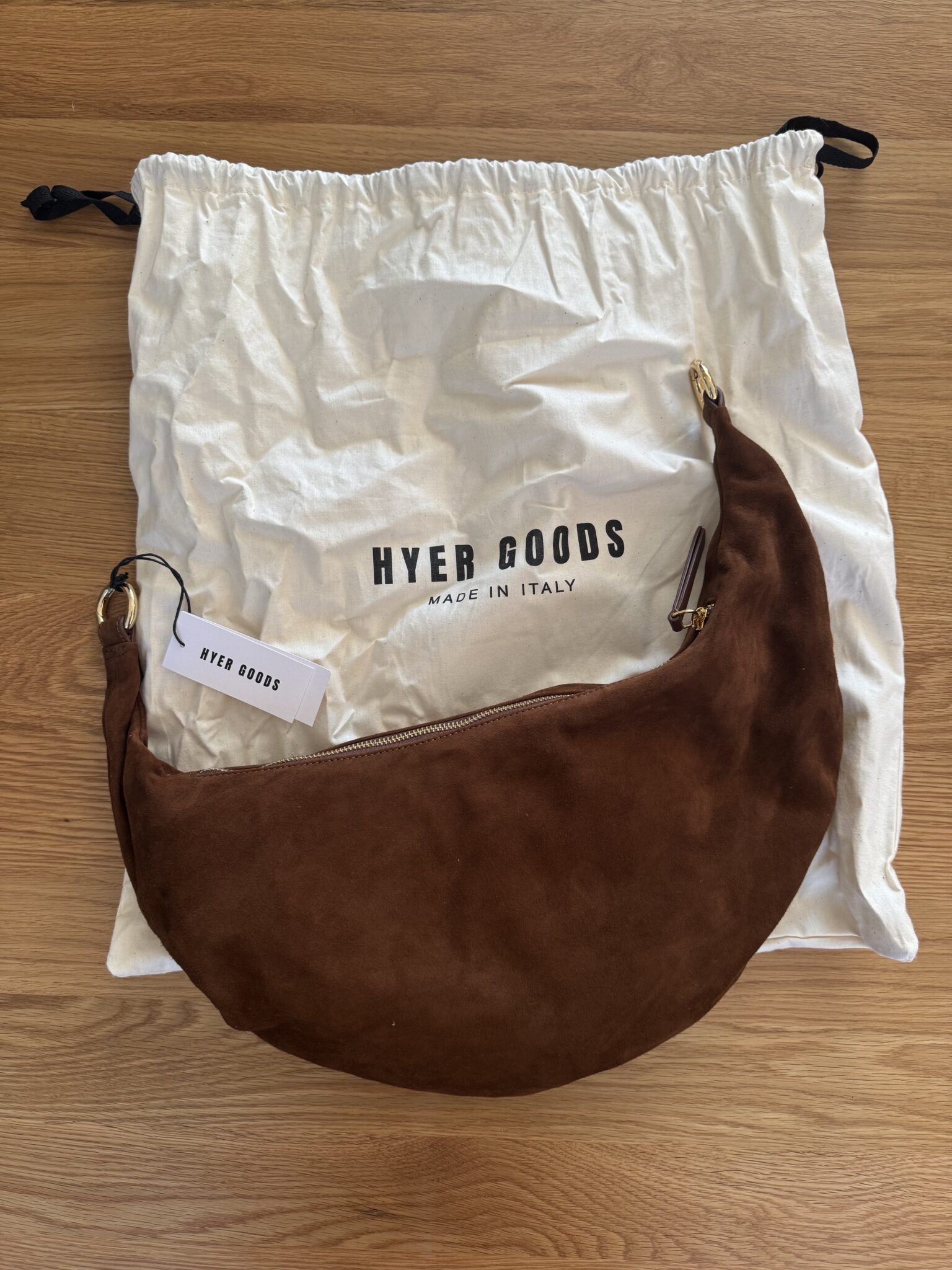A brown crescent-shaped suede bag with a zipper and gold hardware is placed on a white HYER GOODS dust bag on a wooden surface.