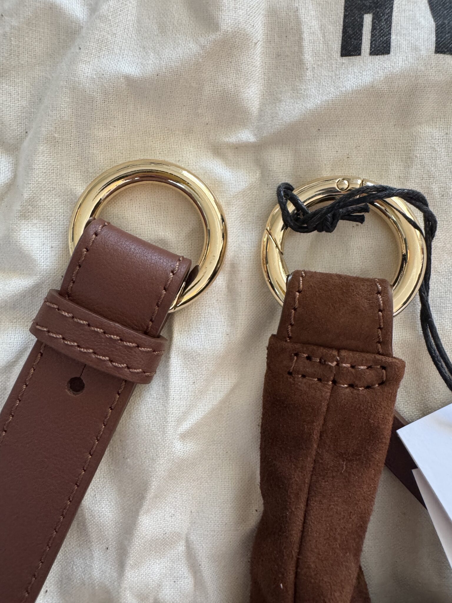 Two brown straps with gold rings are displayed side by side; the left strap is smooth leather, and the right strap is suede with a black string and white tag attached.