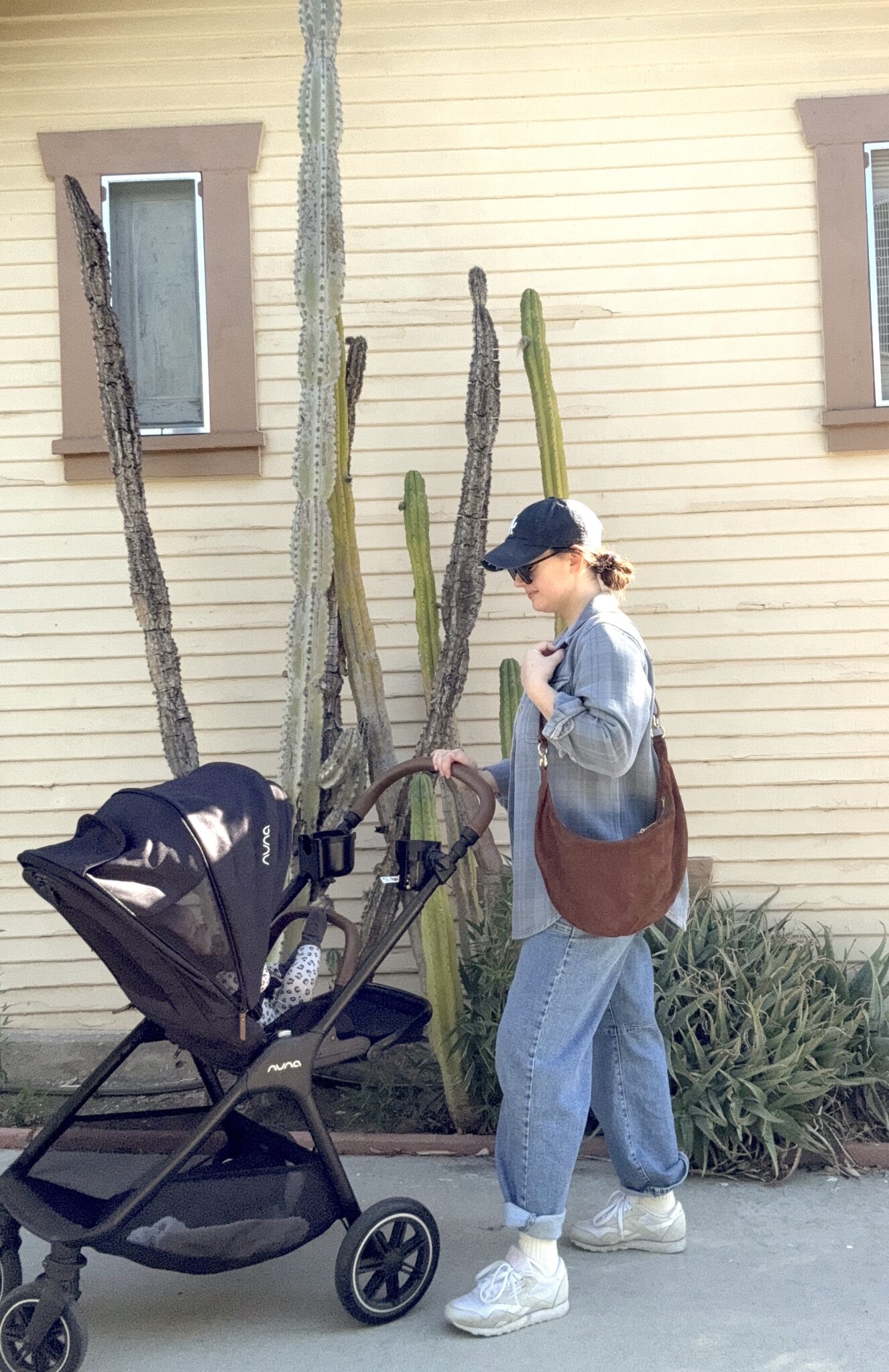 A woman wearing jeans, a cap, and sunglasses pushes a stroller with a baby in front of a beige house with cacti and two windows.