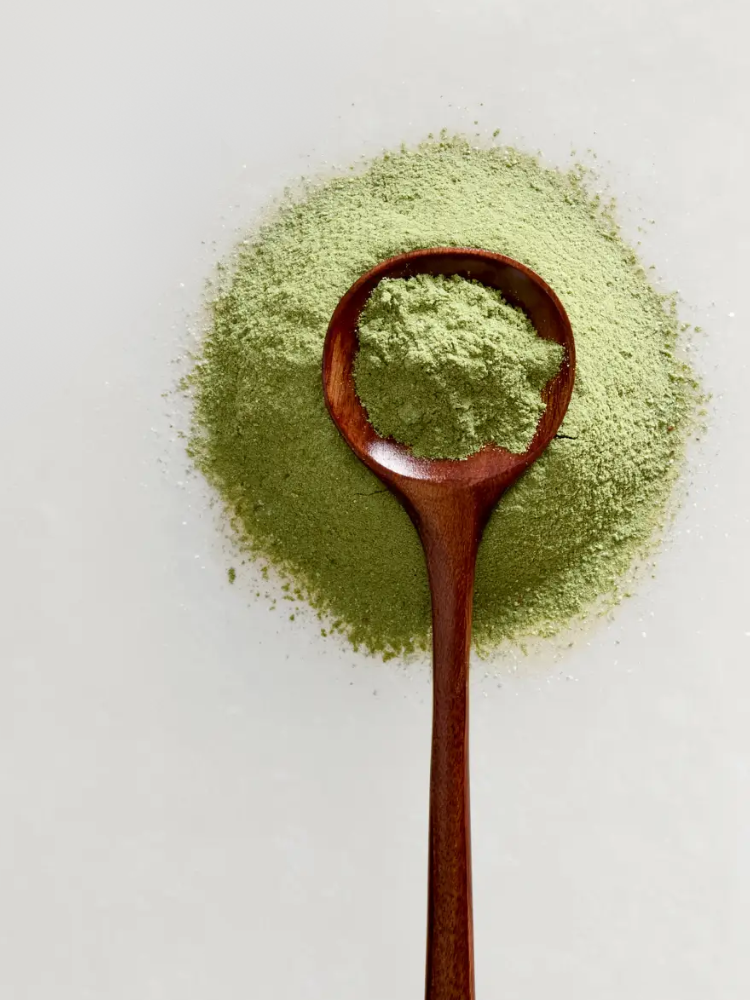 A wooden spoon filled with green powder rests on a pile of the same green powder against a plain white background.