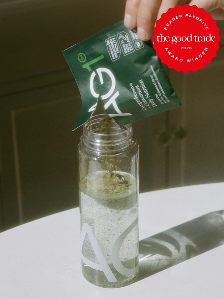 A person pours AG1 supplement powder from a packet into a clear bottle on a table. There's a "Reader Favorite" award badge from "the good trade" in the top right corner.