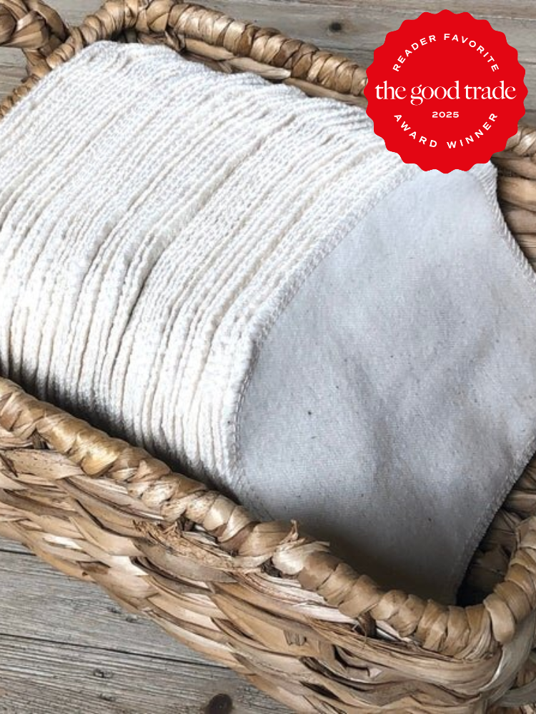 A basket filled with neatly folded, off-white towels on a wooden surface. A red "The Good Trade Reader Favorite Award Winner 2025" seal is at the top right corner.