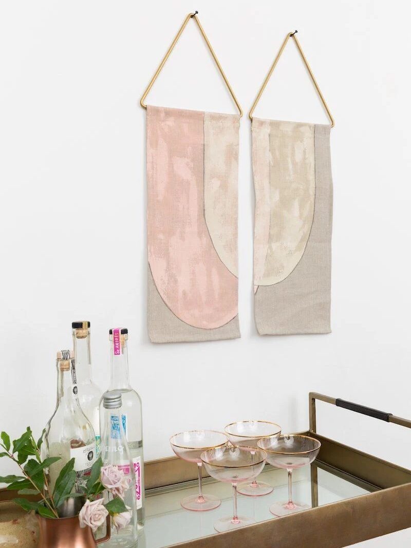 A pink, beige, and grey wall hanging from Conejo & Co placed above a bar cart. 